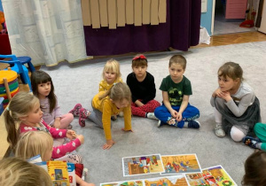 Biblioterapia w grupie 1. Dzieci siedzą w kole, słuchają opowiadania. Na dywanie rozłożone są ilustracje przedstawiające treść opowiadania.